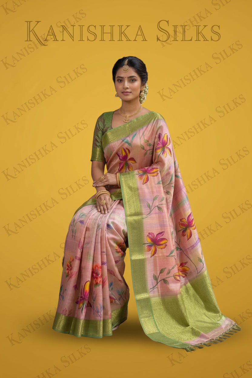 A woman models a pink and green floral printed saree in an outdoor corridor with stone pillars. The saree features a light pink body with vibrant green borders, floral motifs in pink, yellow, and orange, and a green blouse with gold embroidery.