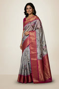 A woman wearing a light gray and pink floral saree with a wide metallic gold border, standing in a traditional courtyard with stone pillars.