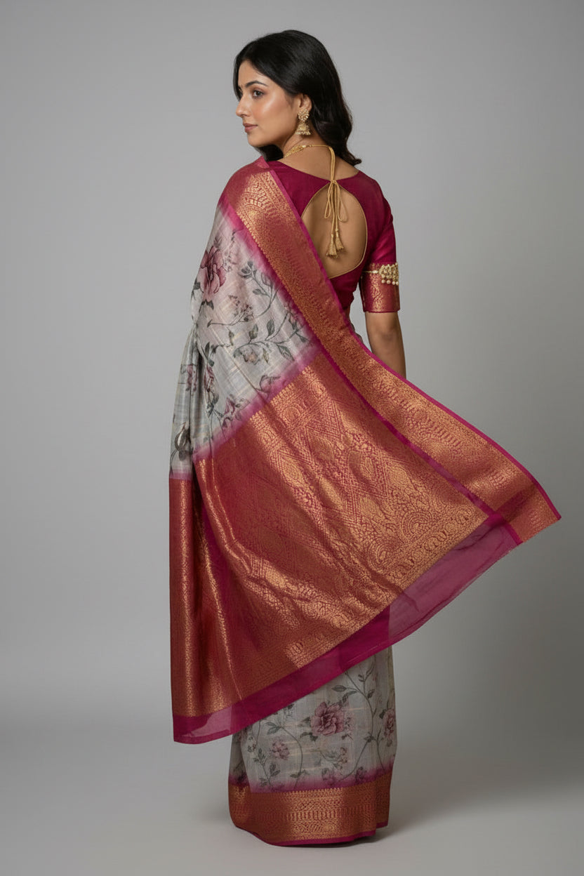 A woman wearing a light gray and pink floral saree with a wide metallic gold border, standing in a traditional courtyard with stone pillars.