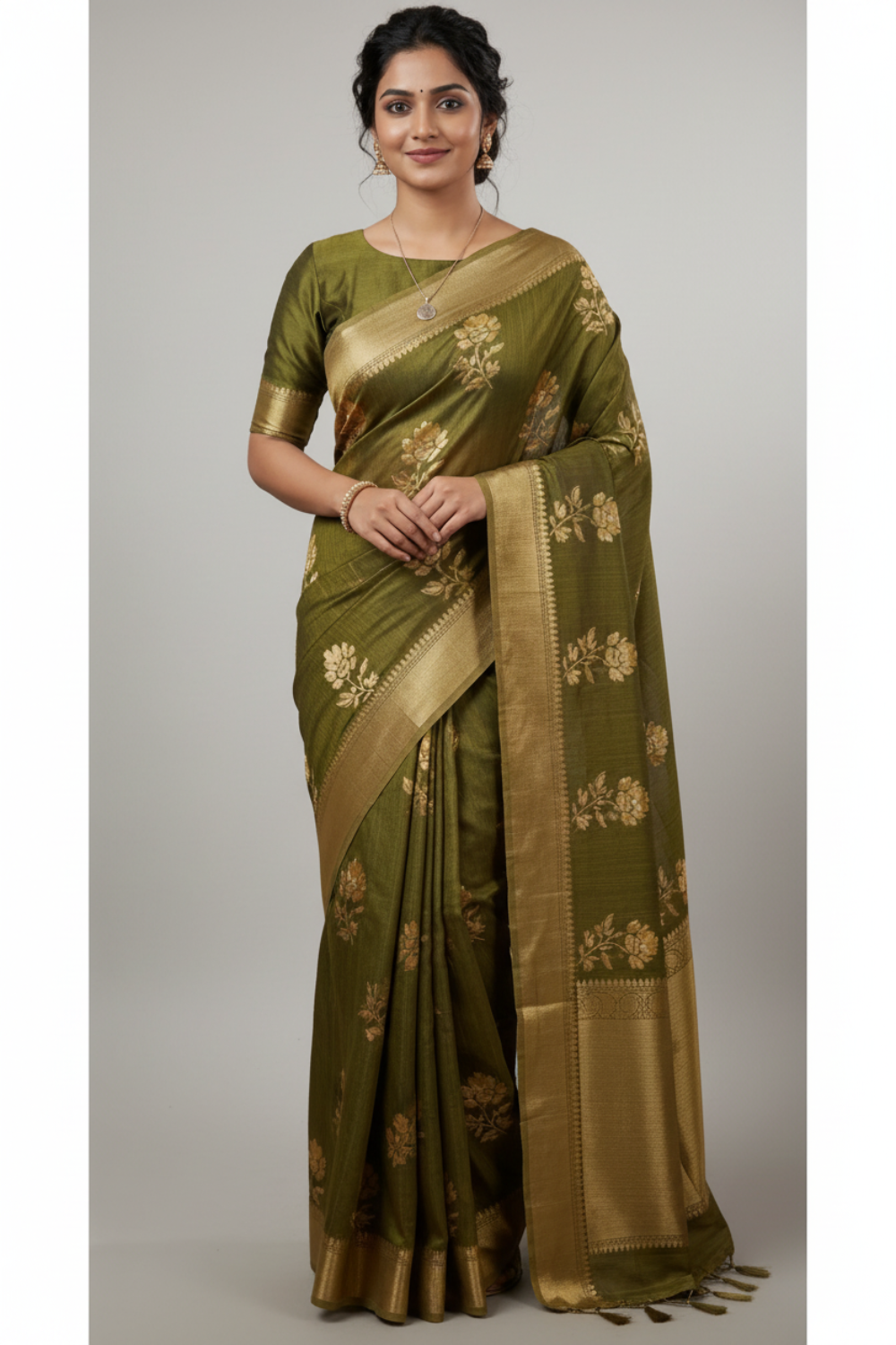 A woman modeling an olive green silk saree with white floral motifs and a gold zari border, standing in a boutique setting with shelves of folded fabrics in the background.